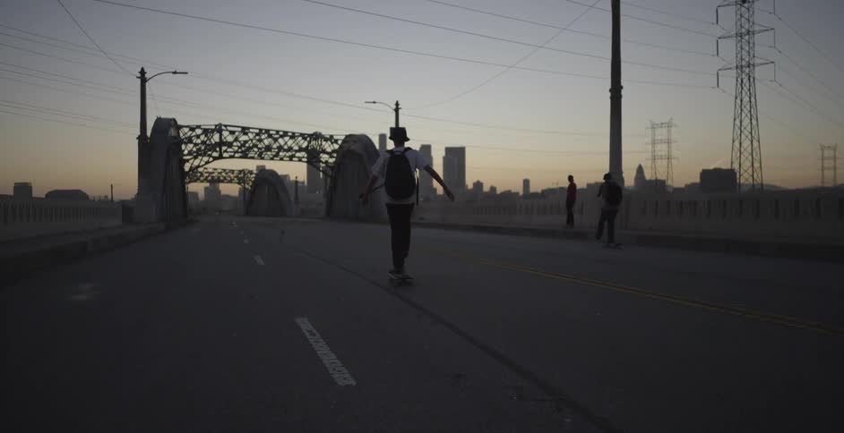A group of teenage boys skateboard across an urban bridge at dawn