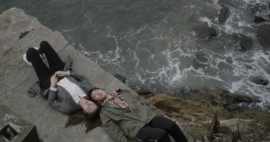 A man and woman lying down on a stone wall above the ocean