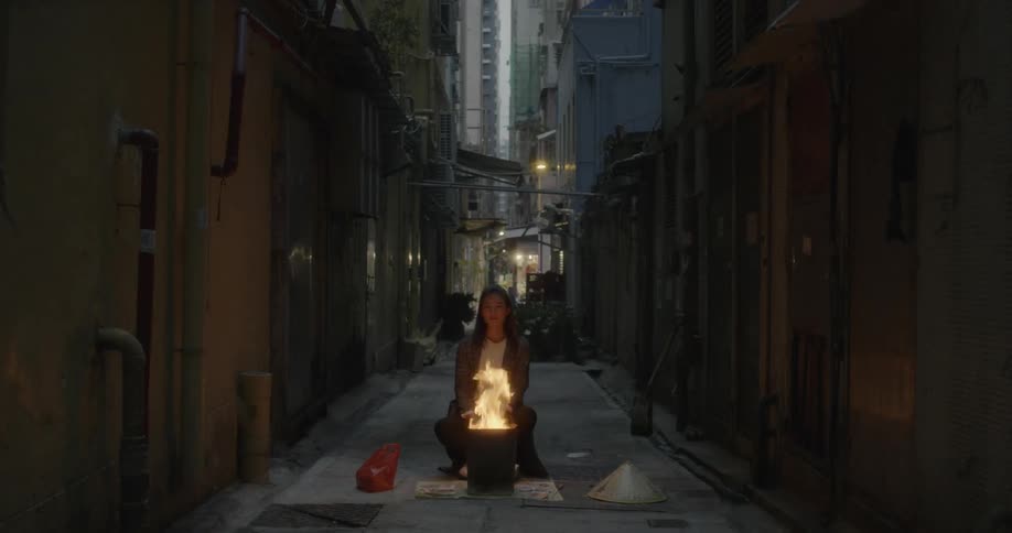 A woman sits by a trashcan fire in a city alley