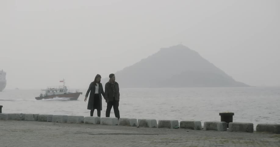 A man and woman hold hands walking along the pier as a boat passes