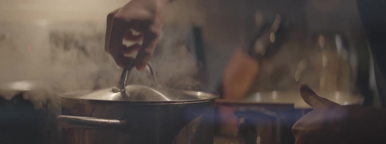 Man lifts lid on steaming pot in kitchen while woman nuzzles his neck