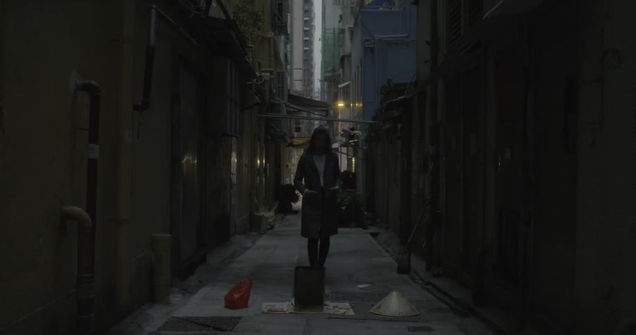 A woman squats down in front of a bin and debris in an alley