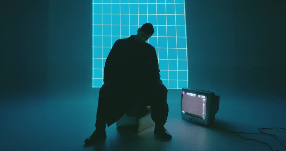 A man sits in a dark room with a tv on the floor and a grid projection illuminating the wall