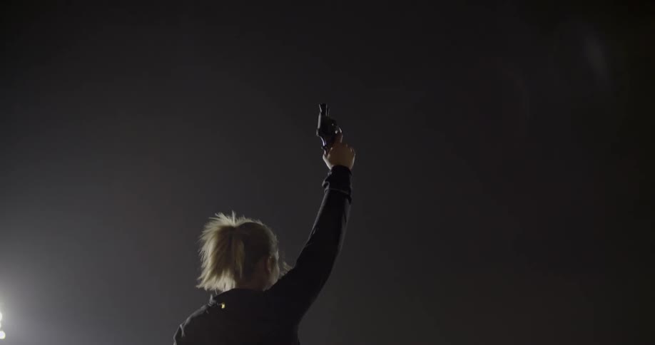 A woman is raises a gun and shoots it into the air as she stands outside