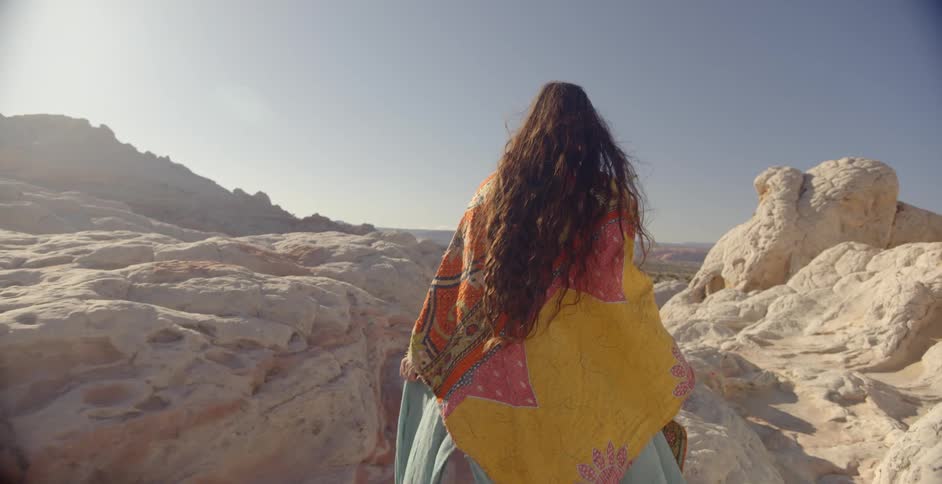 A woman wearing a shawl and dress walks on rocks in the desert