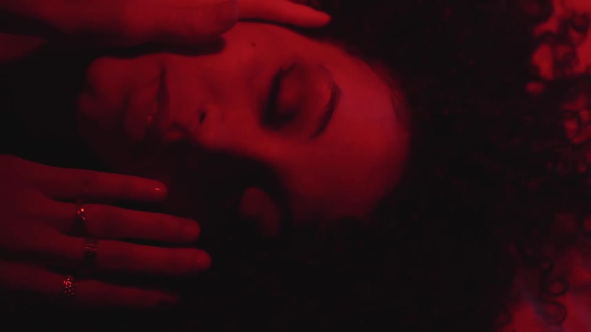 A woman lying on a bed caresses her face and is caressed by another woman
