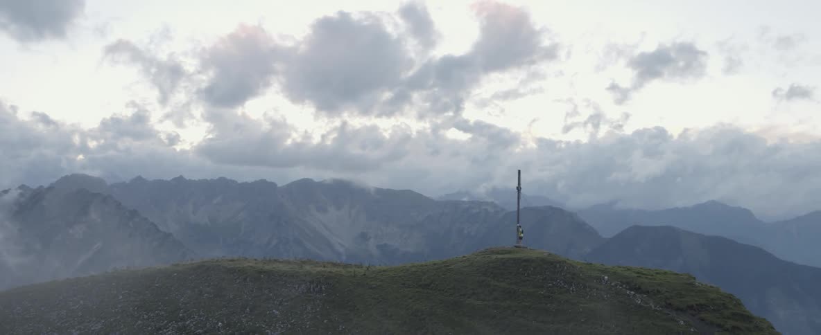 A hiker stands by a metal cross standing on a mountain peak