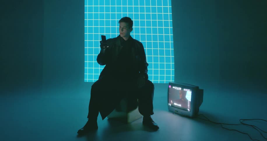 A man sits on a box near a television checking his phone