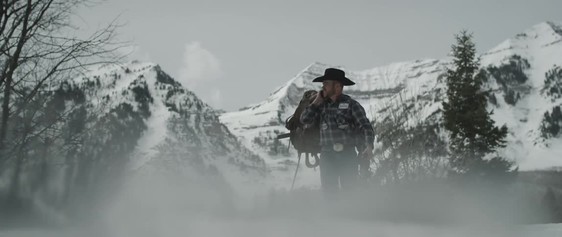 Cowboy carrying a saddle and reins in front of snow covered mountains