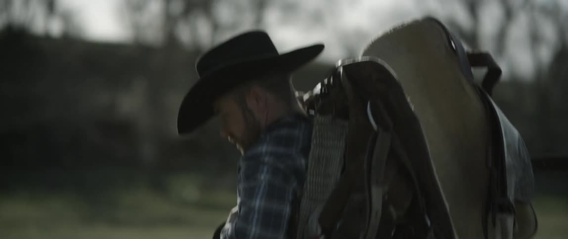 A cowboy carrying a saddle and bridle turns around