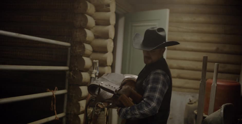 A cowboy carries a saddle in a stable