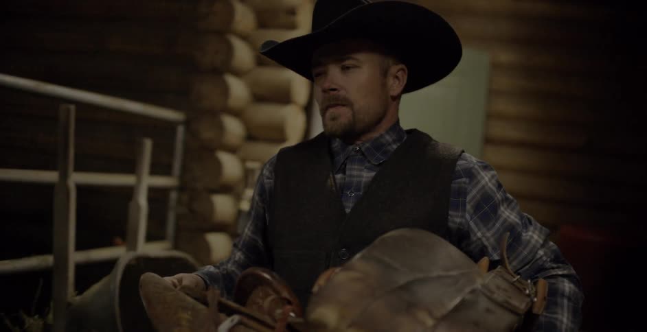 A cowboy carries a saddle in a barn