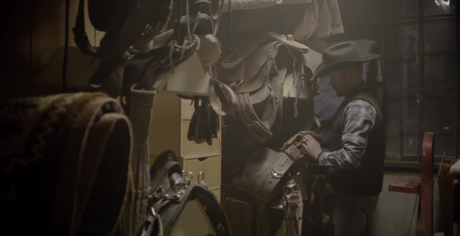 Man wearing cowboy hat picks up and takes saddle from wooden stand