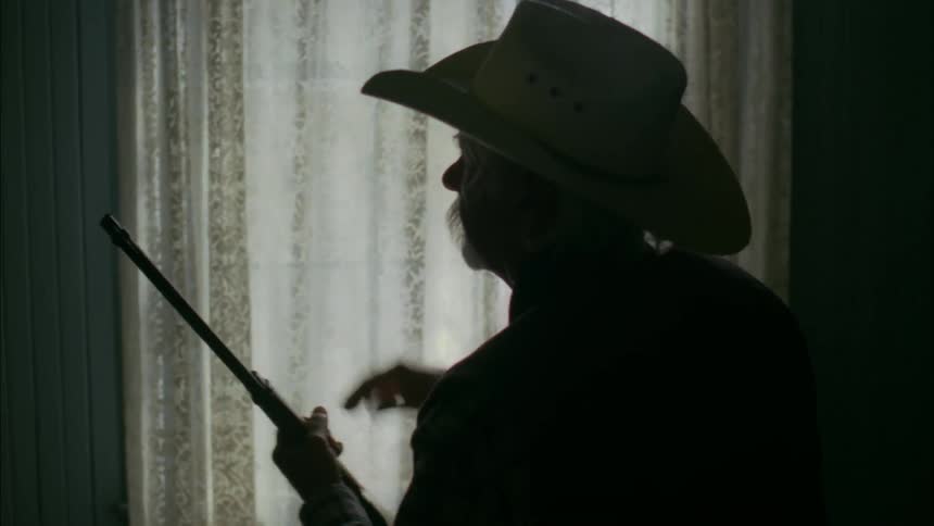 An old man is sitting on the edge of a bed examining a rifle