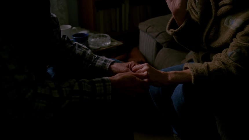 An older couple hold hands in a living room while the woman enjoys a smoke