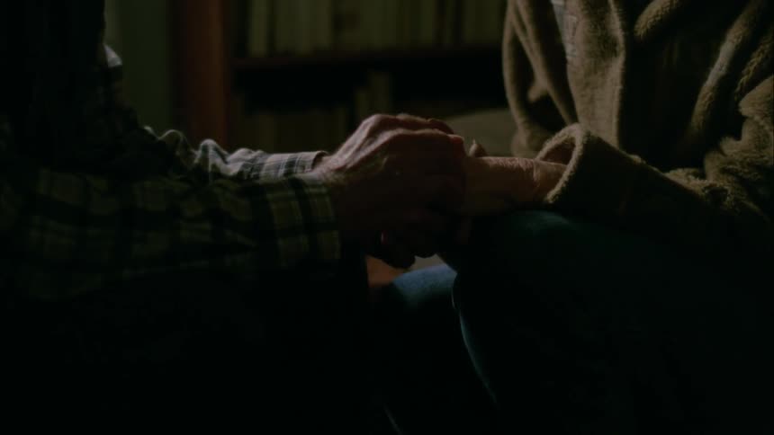 An elderly man sits holding and rubbing an elderly woman's hand