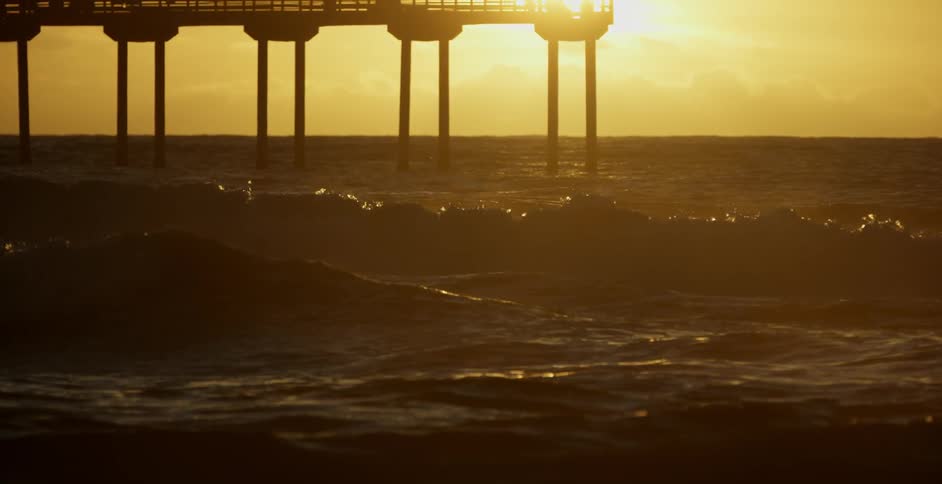 Waves roll towards the shore past a tall pier at sunset