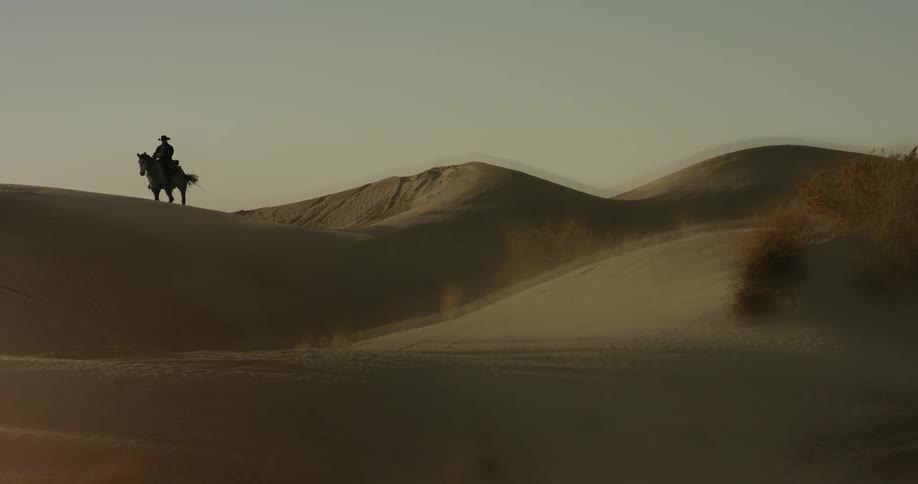 A cowboy is riding a horse over sand dunes during the day