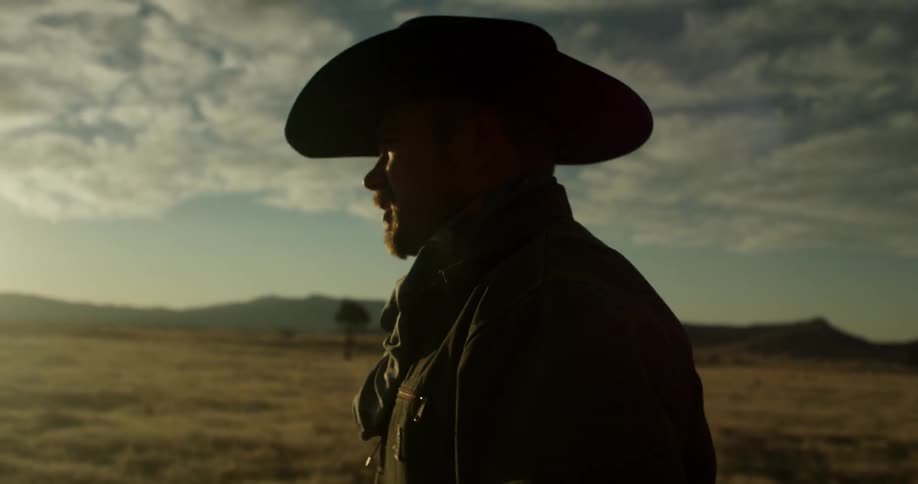 A cowboy walks in a rural field with clouds floating overhead