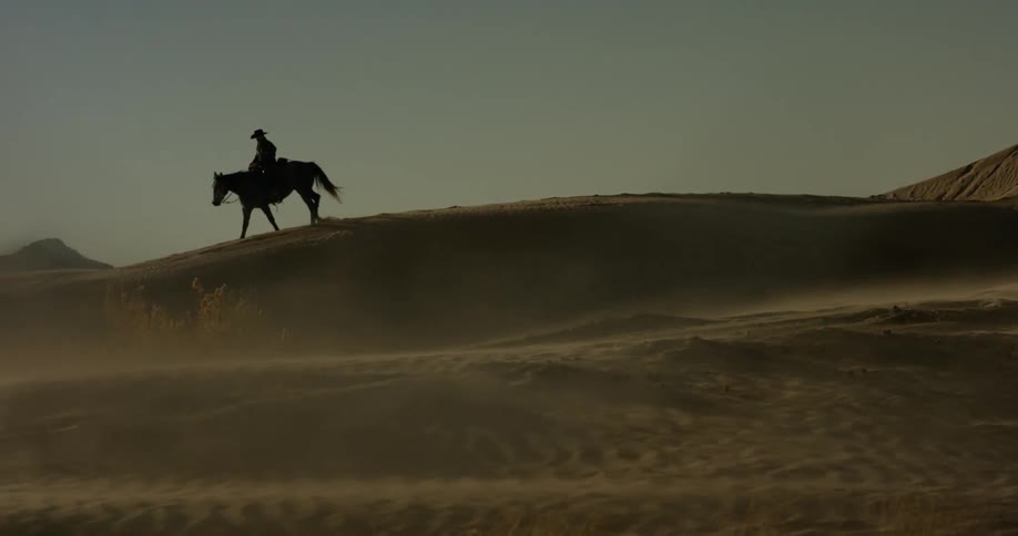 Sand blows across dunes as a man rides a horse over them