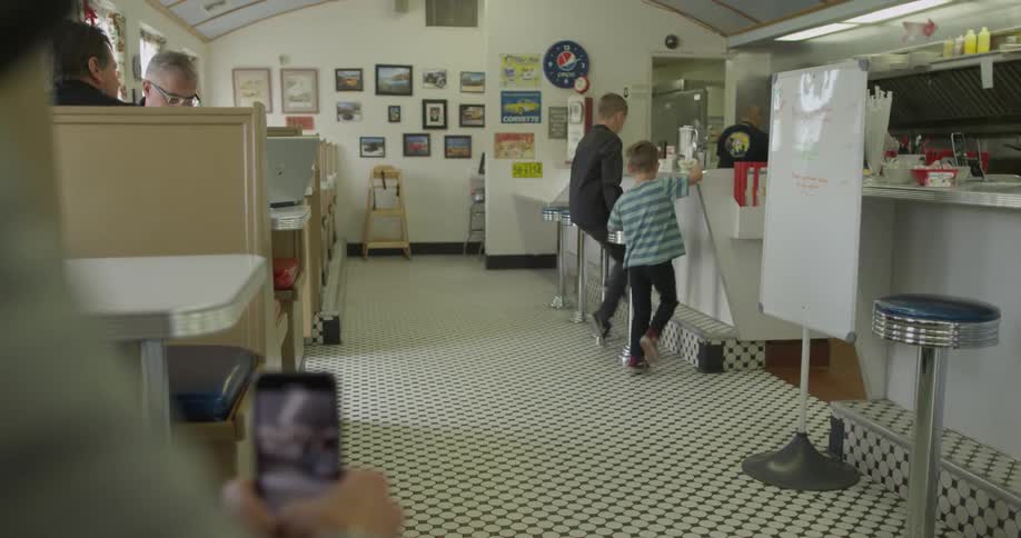 A man films two boys as they walk up and take seats at a diner counter