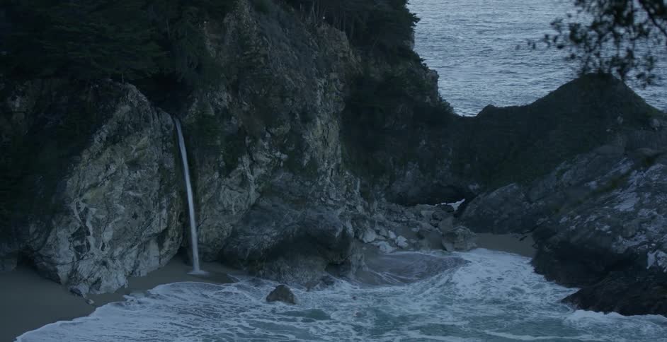 A thin waterfall falls from a cliff onto a sandy shore