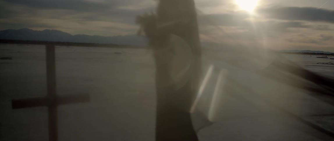 A woman holds flowers as she stands near a cross and a body of water