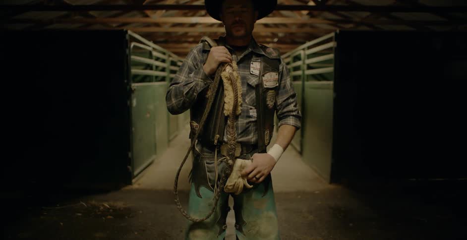 A cowboy stands in a stable with reins over his shoulder