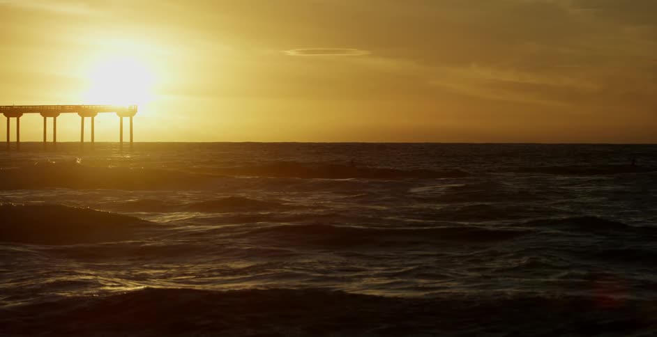 Waves roll towards the shore past a tall pier at sunset