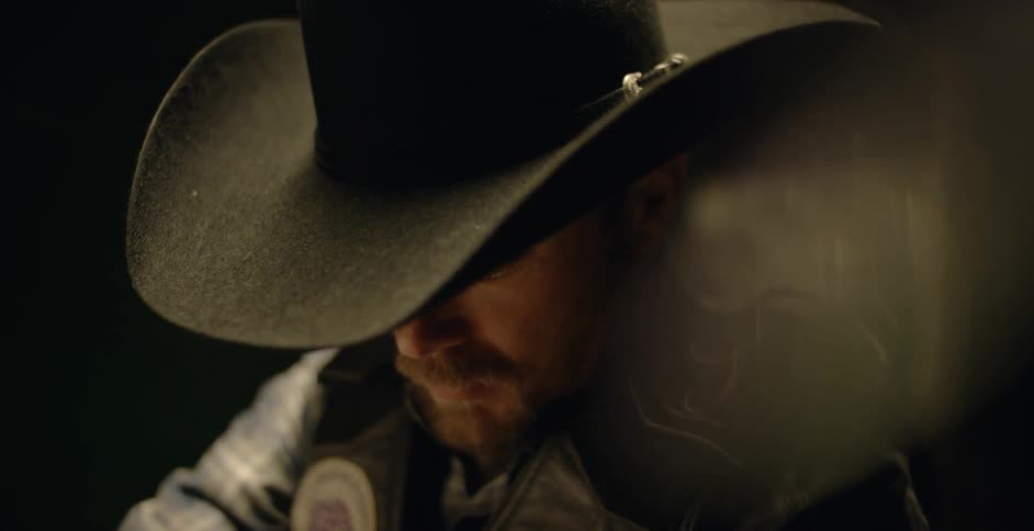 A man wearing a cowboy hat sitting on something and looking down