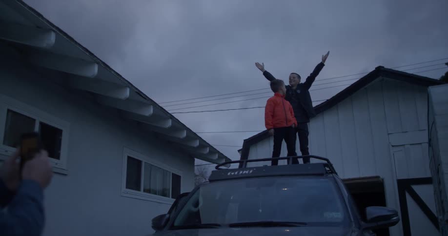 A person is taking a picture of two kids standing on the top of a car