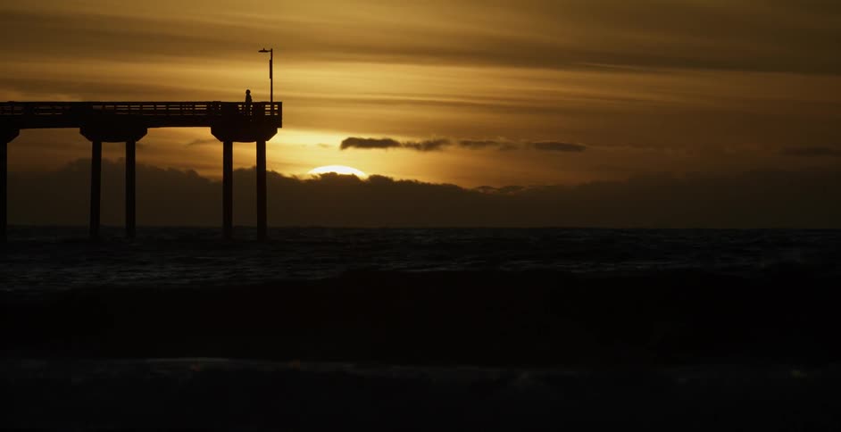 A person is walking near the end of a tall pier at sunset