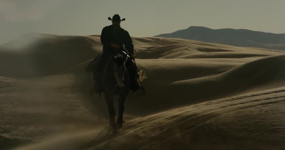 Man wearing cowboy hat riding horse over sand dunes while wind blows