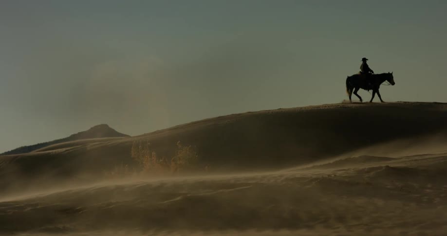 Person rides a horse in a desert with the dust blowing at sunset