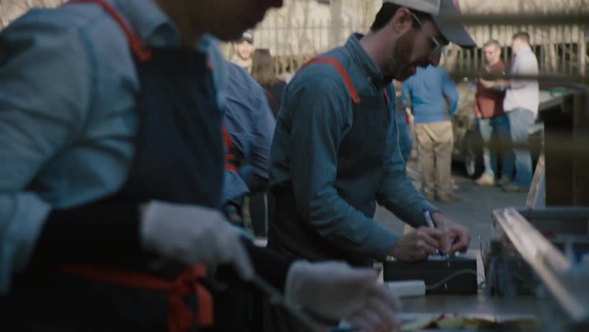 Man spreads sauce on uncooked pizza dough outside at a table