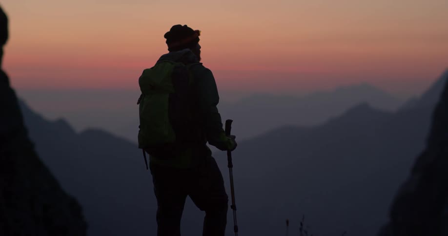 A hiker stops on a mountain ledge and looks out at the sunset