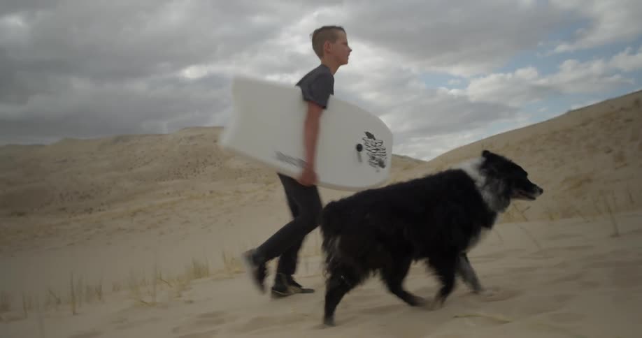 A boy carrying a bodyboard walks up a sand dune with his dog