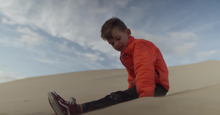 A young boy sits on a sand dune and pushes his hands in the sand