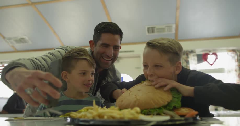 A man records a kid taking a bite of a huge burger