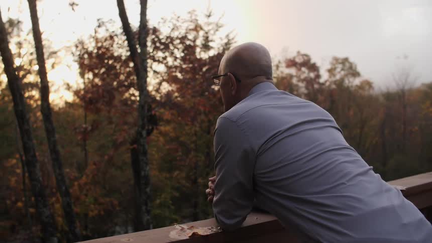 A man leans on a balcony railing looking out towards trees
