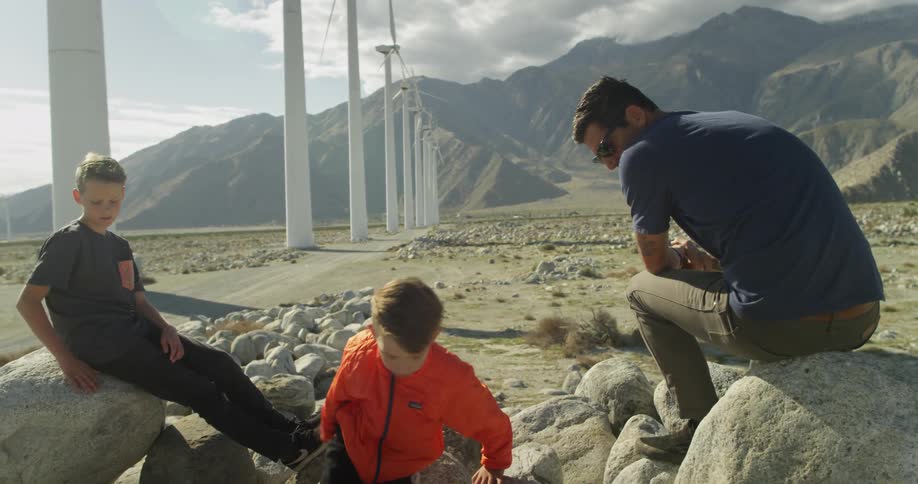 A man and two children sit on some rocks by windmills