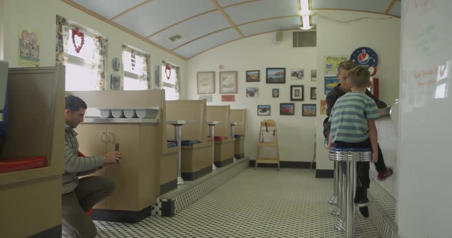 Two children sitting on stools in a diner while a man records them with a phone