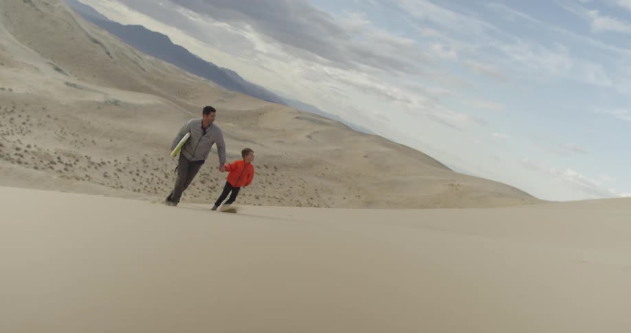 A man holds his son's hand as they walk up a sand dune