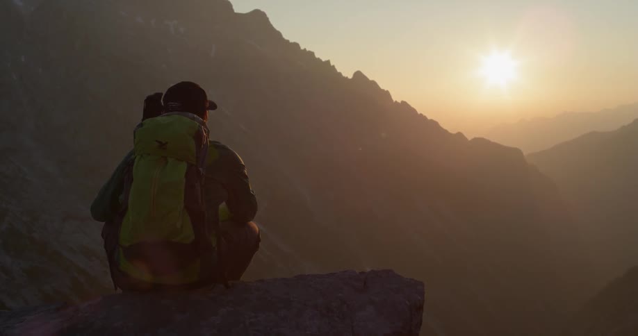 A hiker sits atop a cliff looking towards the sun setting