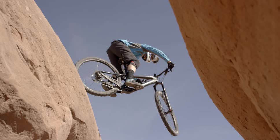A person jumping between two large rocks on a bike while being filmed underneath