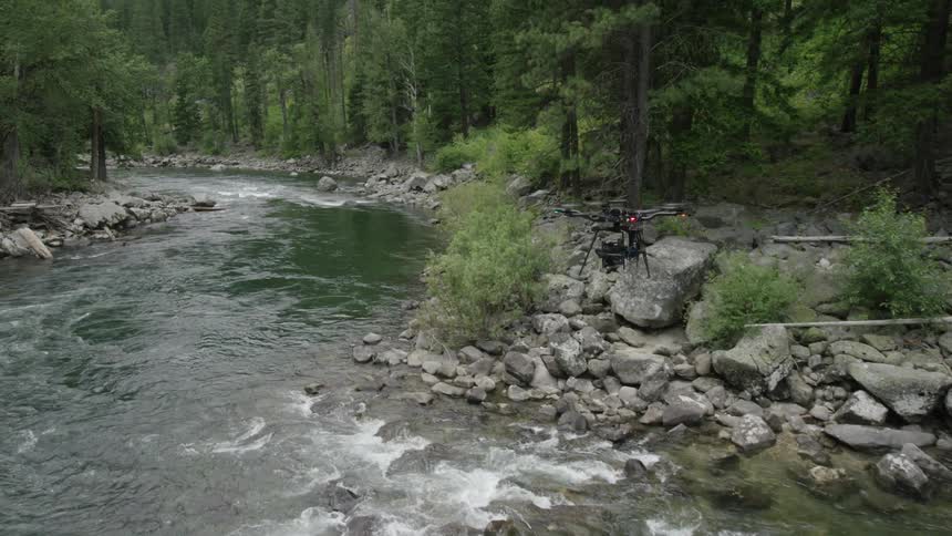 A drone flies over a rocky river in a forest during the day
