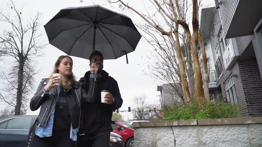 A man and woman hold coffee cups and walk under an umbrella
