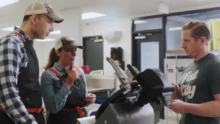 People stand and talk near a meat slicer in a large kitchen during the day