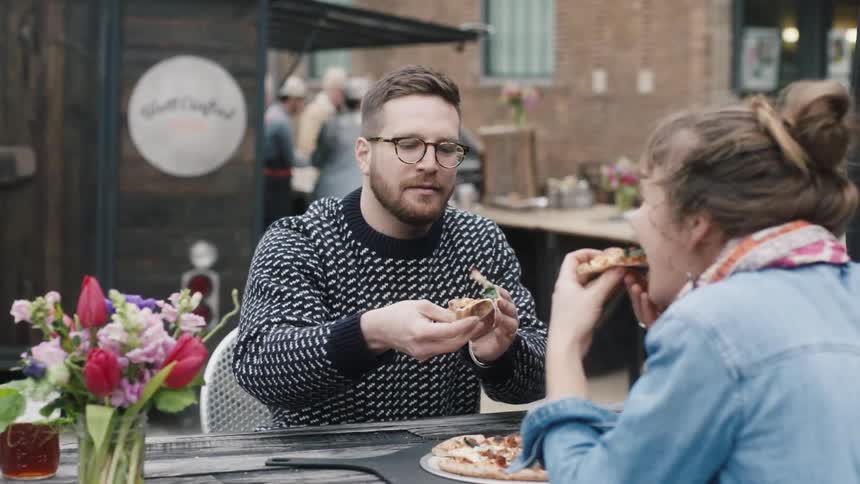 A man and woman eat pizza outdoors at a table