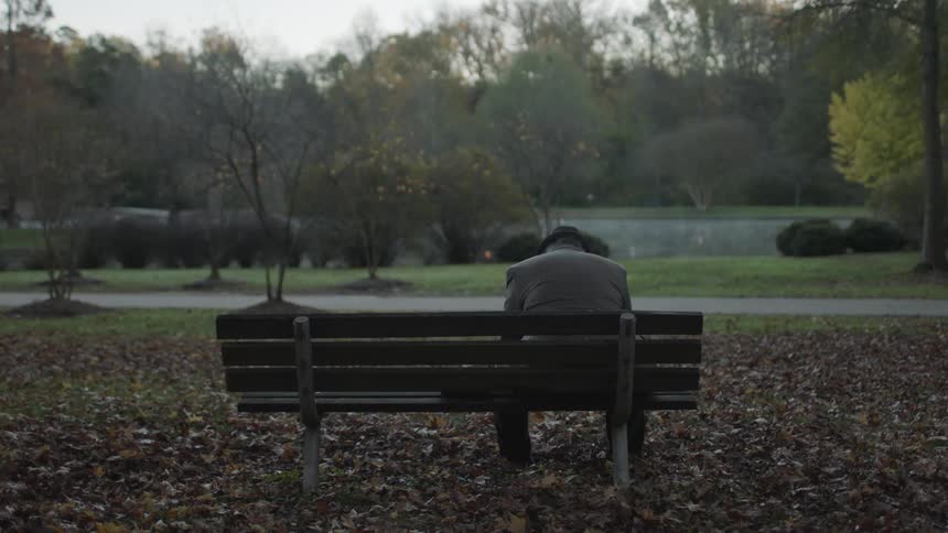 An older man sitting on a park bench throws leaves dejectedly